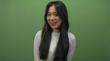 İzole edilmiş yeşil bir arka plan duvarına gülümseyen kadın beyaz bir gömlek ve uzun siyah saçlı genç Çin zarafeti ve modern gündelik moda somutlaştırılmış.