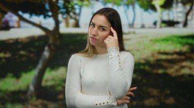 Güneşli bir parkta ağaçlarla çevrili bir yerde beyaz bir gömlekle gezen ve poz veren düşünceli bir kadın.