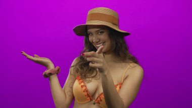 Hasır şapkalı ve turuncu bikinili kadın pembe stüdyoda kameraya parmak ucuyla bakıyor..