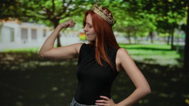 Güneşli parkta kaslarını kasan bir kadın ağaçlardan ve yeşillikten oluşan bir zemin ile güç ve kendine güven gösterisi yaparak güçlü ve güçlendirici bir açık hava manzarası yaratıyor..