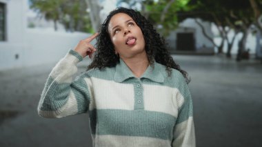 Kıvırcık saçlı bir kadın, sakin bir sokakta anlamlı jestler yapıyor. Duygularını, barışçıl bir arka plana karşı canlı bir şekilde sergiliyor..