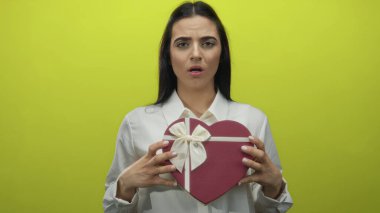 Kadının elinde kırmızı kalp şeklinde bir hediye kutusu var. Parlak sarı bir zemin üzerinde. Yüzünde belirsiz bir ifade var..