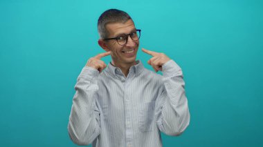 Gözlüklü ve çizgili gömlekli bir adam mavi arka planda gülüşünü gösteriyor, izole edilmiş bir stüdyoda güven ve neşe yayıyor..