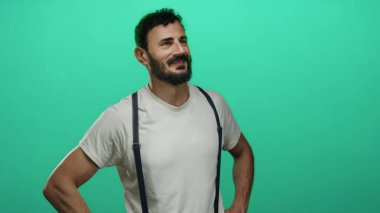 Sakallı İspanyol sakallı, elleri kalçasında canlı yeşil bir arka plana yaslanmış bir şekilde gülümseyen, stüdyo ortamında günlük beyaz gömlek ve pantolon askısı takan bir adam.