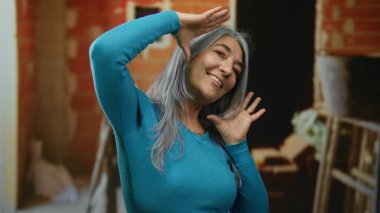 Gri saçlı, gülümseyen ve kapalı bir inşaat alanında neşeli poz veren yaşlı bir kadın kısmen inşa edilmiş tuğla bir duvarın arka planına karşı mutluluk ve enerji sergiliyor..