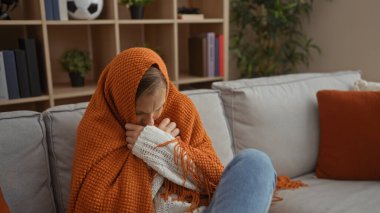 Turuncu bir battaniyeye sarılı bir kanepede oturan kadın, üşümüş, kitap ve bitkilerle dolu sıcak bir oturma odasında, evin içinde sıcaklık ve rahatlığı öneriyor..