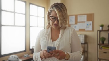 Güneş ışığıyla aydınlanmış bir ofis odasında, gözlüklü sarışın kadın akıllı telefon tutuyor ve kaşlarını çatıyor..
