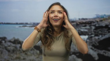 Heyecanlı yüzlü bir kadın bulutlu bir gökyüzünün altında kayalık bir sahil kenarında duruyor. Elleriyle doğanın güzelliğini ve duygularını gösteriyor..