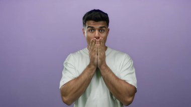 Genç adam, canlı bir mor arkaplan üzerine izole edilmiş, sıradan beyaz bir gömlek giyen, yüzün yanında elleri olan bir dizi duygu sergileyen, şaşırdığını ifade ediyor..