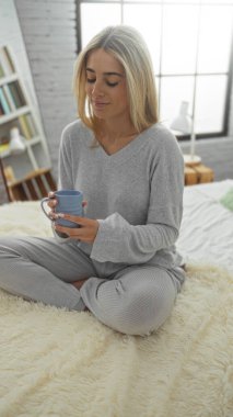 Yatak odasında rahat bir şekilde elinde bardakla rahat bir pijama giyen, camdan içeri süzülen yumuşak kumaşlı, dingin bir atmosferi olan bir kadın..