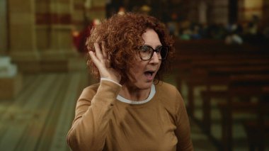 Kıvırcık saçlı ve gözlüklü bir kadın loş bir kilisede kahverengi bir gömlek ve el kulağına bir jest giyerek şaşkınlığını ifade ediyor..