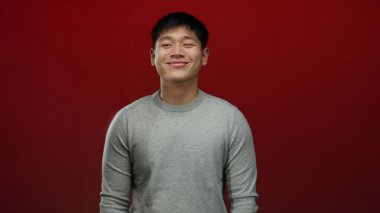 Gri süveter giymiş genç Çinli bir adam izole edilmiş kırmızı bir arka plan karşısında gülümsüyor. Mutluluk ve basitliği ifade ediyor..