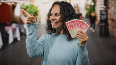 Restoran terasında gülümseyen bir kadın, elinde İsrail şekeri, parmakla gösterişli süveter giyerek para gösteriyor..