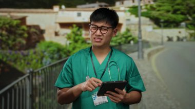 Tıbbi önlüklü genç adam şehir caddesinde tablet kullanıyor, boynunda steteskop ve yüzüne yansıyan güneş ışığıyla dikkatle odaklanıyor..