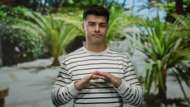 Genç adam parmaklarıyla bir açık hava parkı ortamında, yemyeşil ile çevrili, çizgili bir kazak giyiyor, oyuncu ve kendinden emin bir tavır sergiliyor..