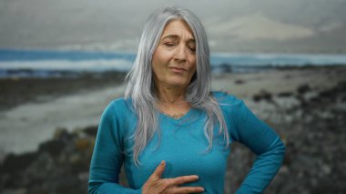 Mavi tişörtlü yaşlı bir kadın kumsalda karnını tutuyor bulutlu bir gökyüzünün altında kıyı arka planına karşı iyi görünmüyor..