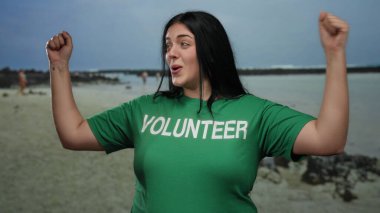 Deniz kıyısında yeşil gönüllü gömleğiyle gülümseyen bir kadın, heyecan ve coşku gösteriyor, arkada açık mavi gökyüzünün altında okyanus dalgaları var..