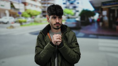 Genç İspanyol bir adam şehrin bir caddesinde duruyor, kışlık bir ceket giyiyor, soğuk ama yakışıklı görünüyor, açık hava ortamında elleri sımsıkı bağlı..