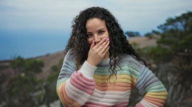 Sahilde renkli bir süveter giyen, deniz manzaralı bir kadın gündüz vakti neşeli bir hava sergiliyor..