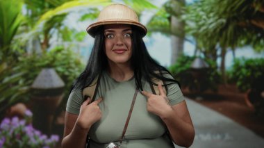 Şaşırmış ifadesi olan bir kadın turist, tropikal bir şapka takmış ve elinde antika bir kamerayla kendini açık hava parkında gösteriyor..