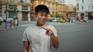 Şehir caddesinde ihtiyatlı bir jest yapan genç adam, arka planda binalar ve bulanık yayalarla canlı şehir yaşamını ve kültürel çeşitliliği vurguluyor..