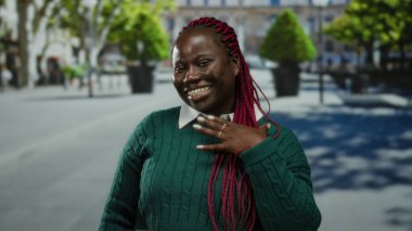 Arka planda ağaçlarla dolu, açık havada gülümseyen bir kadın canlı saçlarını ve yeşil süveterini güneşli bir ortamda neşeli bir şehir havası yayarken gösteriyor..