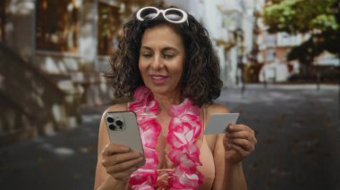Hawaii zambağı olan bikinili bir kadın sokakta akıllı telefon ve kredi kartı kullanıyor. Şehir ortamında mutlu ve rahat görünüyor..