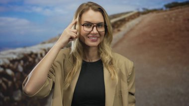 Açık havada gülümseyen bir kadın kafasında parmağıyla düşünceli bir jest yapıyor. Sarı saçlar günlük güneşlik, mavi gökyüzü gözlükleri arka planda doğa ilhamı kavramı.