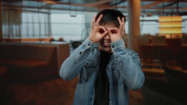 Genç Çinli bir adam gözlerinde gözleriyle gözlük jesti yapıyor, sıradan bir kot ceket giyiyor, modern dekorlu ve çevre aydınlatmalı rahat bir kahve dükkanında gülümsüyor..