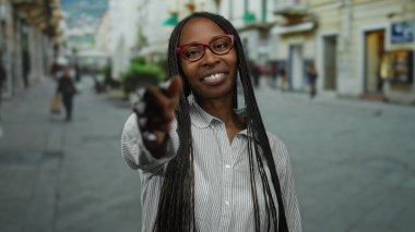 Şehirdeki sokakta el sıkışmak için elini uzatan kadın, şehir arka planında bulanık, açık havada dostça bir etkileşime işaret ediyor..