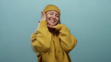 Sade sarı bir kazak ve uyumlu bir bereyle gülümseyen bir kadın, saf mavi bir arka planda sıcaklığı ve mutluluğu izole bir duvarda yakalıyor..