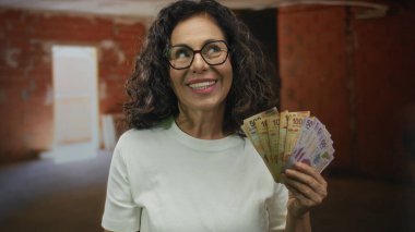 İnşaat alanında elinde Meksika pezosu tutan ve susma hareketi yapan bir kadın, gizliliği vurgulayarak, orta yaşlı, İspanyol, nakit, finans, iş yeri, gözlük, gülümseme.