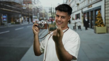 Elinde klasik kamera mimikleri olan genç bir adam açık şehir ortamında gündüz vakti işlek şehir caddesinde davetkar bir şekilde el kol hareketi yapıyor.