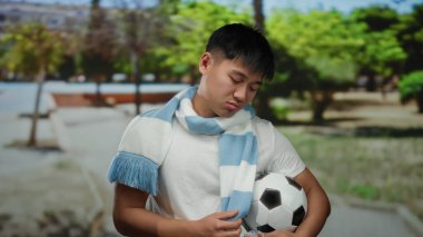 Parkta elinde futbol topuyla gezen ve eşarp takan genç bir adam, güneşli bir açık havada uykulu görünüyor, arka planda yeşil ağaçlar var..