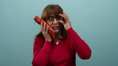 Kırmızı gömlekli kadın, mavi arka planda yüzüne buz torbası dayayarak acı çekiyor. Rahatsızlığı vurguluyor ve yardım istiyor..