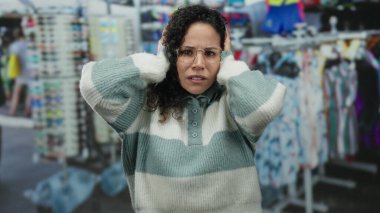 Hırkalı bir kadın yoğun bir cadde pazarında renkli giysiler ve aksesuarlarla boğulmuş durumda. Hayal kırıklığı ve kafa karışıklığını ifade ediyor..