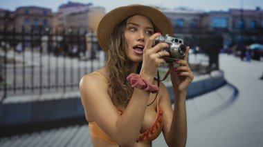 Bikini giymiş bir kadın elinde kamera, hareketli bir caddede duruyor, açık hava sahnelerini ilginç bir ifadeyle yakalıyor, modayı keşif ve gençlik ile harmanlıyor..
