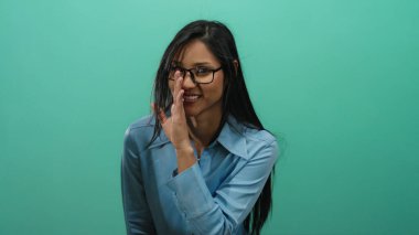 Canlı yeşil bir arka plana karşı neşeli bir ifadeyle fısıldayan, gözlüklü ve mavi gömlekli bir kadın iletişim temaları için çekici ve ulaşılabilir bir görsel yaratıyor.