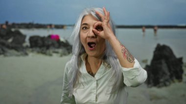 Üst düzey bir kadın, kır saçlı ve neşeli bir ifadeyle manzaralı bir sahil sahilinde dikilip açık havanın tadını çıkarırken, gözünün önünde iyi bir jest yapıyor.