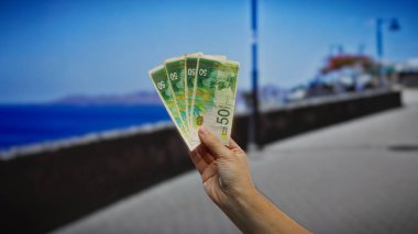 İsrail şekellerini sahil caddesinde tutan adam yaz tatili, para ve yatırım temaları vurguluyor.