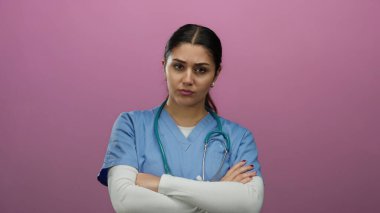 Mavi önlüklü ve steteskoplu İspanyol hemşire kollarını izole edilmiş pembe bir arkaplanda kavuşturarak mesleki güven ve sükunet gösteriyor..