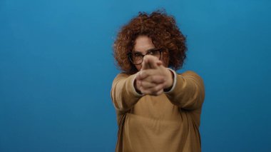 Latin bir kadın, parlak mavi bir duvara karşı silah hareketi yapıyor. Kendine güveni ve mizahı ifade ediyor. Gözlük takıyor ve kahverengi bir süveter giyiyor..