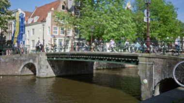 Güneşli bir günde ikonik mimari ve yemyeşil sergilenen, bisikletlerle köprüyü çevreleyen Amsterdam Kanalı 'nın bulanık görüntüsü.