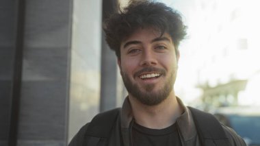 Güneşli bir günde açık havada gülümseyen, sıradan bir ceket ve sırt çantası giyen, yumuşak odaklı arka planı olan bir binanın yanında duran İspanyol asıllı genç bir adam..