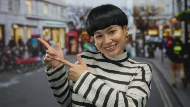 Şehir yaşamının ve dükkanların bulanık görüntüsüyle çevrili hareketli bir şehir caddesini işaret eden genç bir kadın gün ışığında enerjik bir açık hava atmosferini yakalıyor..