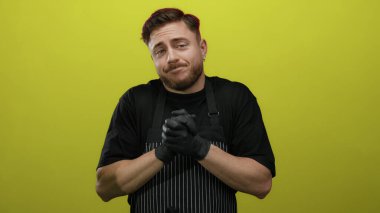 Önlük ve eldiven takan sakallı genç adam parlak sarı arka planda duruyor ve neşeli bir ifadeyle omuz silkiyor..