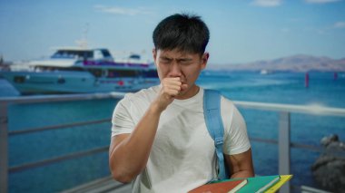 Sırt çantalı genç adam deniz kenarında öksürüyor arka planda teknesi olan kitapları açık gökyüzünün altında açık hava öğrenciliğini simgeliyor..