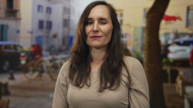 Dolu bir caddede parmakları dudaklarında şşşş yapan bir kadın, kentsel çevre ile çevrili, canlı bir açık hava ve canlı şehir manzarası sergiliyor..