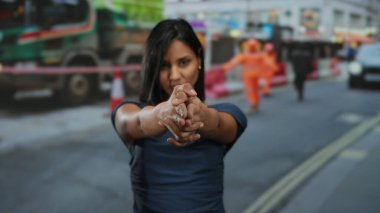 Genç bir kadın, arka planda bulanık işçiler ve araçların bulunduğu bir inşaat alanındaki silaha benzeyen el hareketi yapıyor..