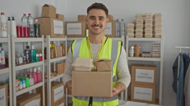 Yansıtıcı yelek giyen genç adam bir hayır kurumunda gönüllü olarak çalışıyor, içeride gıda paketleri düzenliyor, sosyal yardım çabalarını ön plana çıkarıyor..